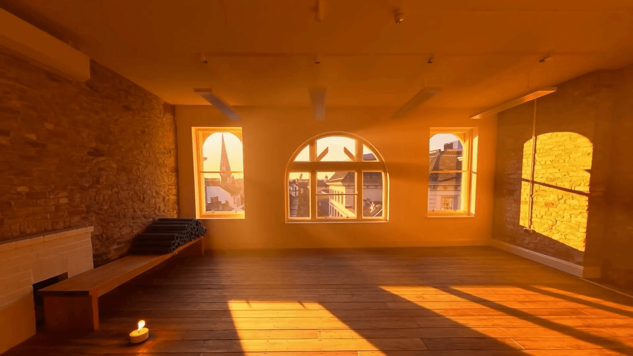 A rolled yoga mat unfurling across a sunlit studio floor, long window shadows on a warm plaster wall.